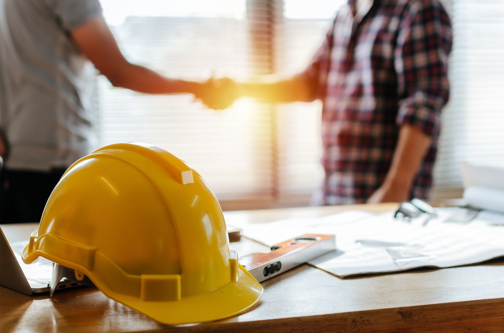 An architect and a construction worker shake hands backlit by sunlight. A construction hat and site plans are on a desk in the foreground.