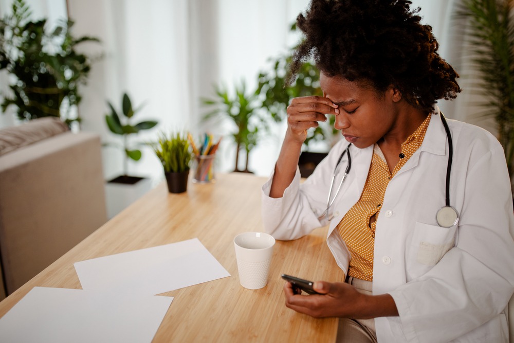 A nurse, sitting at a table, pinching her forehead with eyes closed, holding her phone, unhappy about a license suspension.