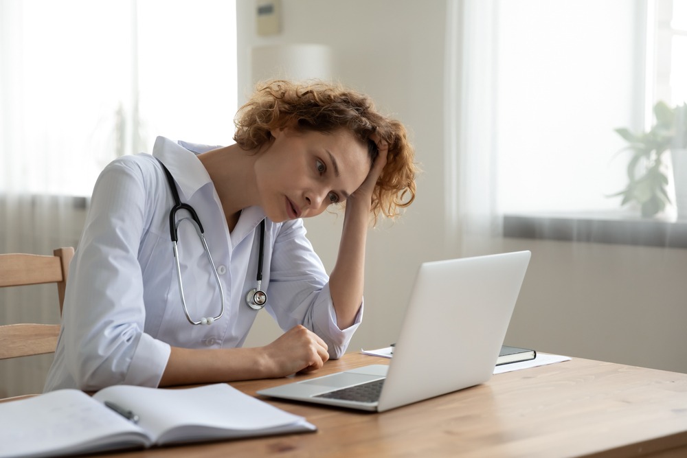 A woman in scrubs with a stethoscope hanging from her neck has her hand in her hair while looking at a computer