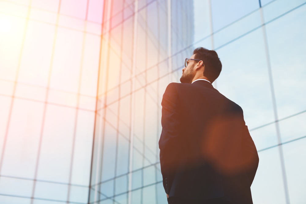 A bearded property tax professional wearing glasses and a suit, facing the light reflecting from a glass-paneled office building.